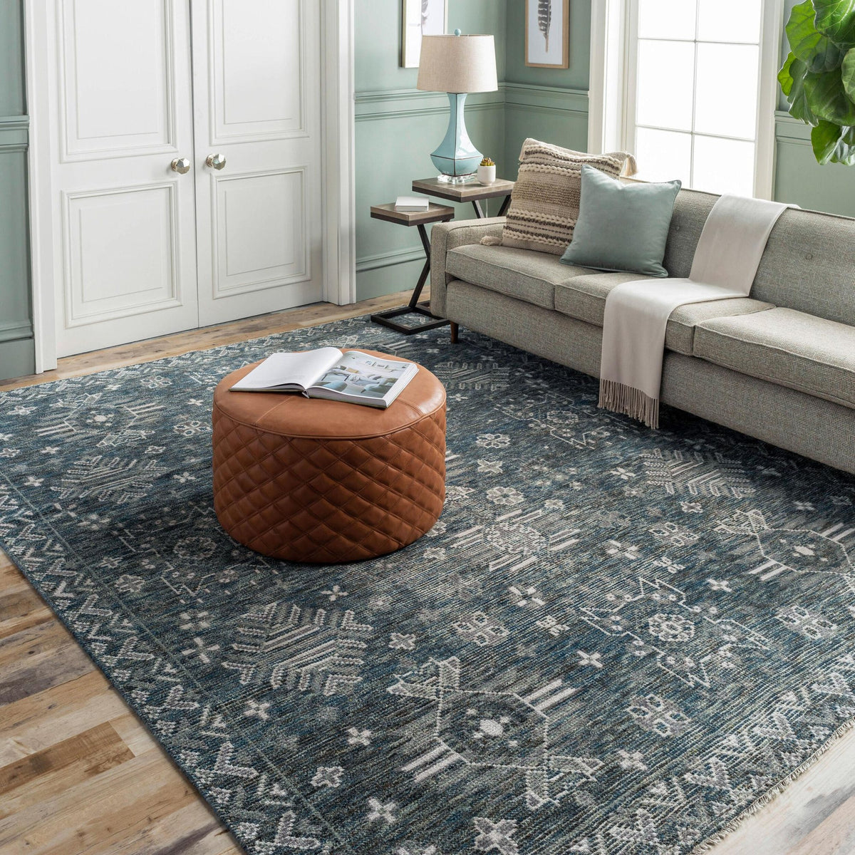 Living room with a blue-grey patterned rug, cognac leather pouf, sofa with pillows and a blue table lamp